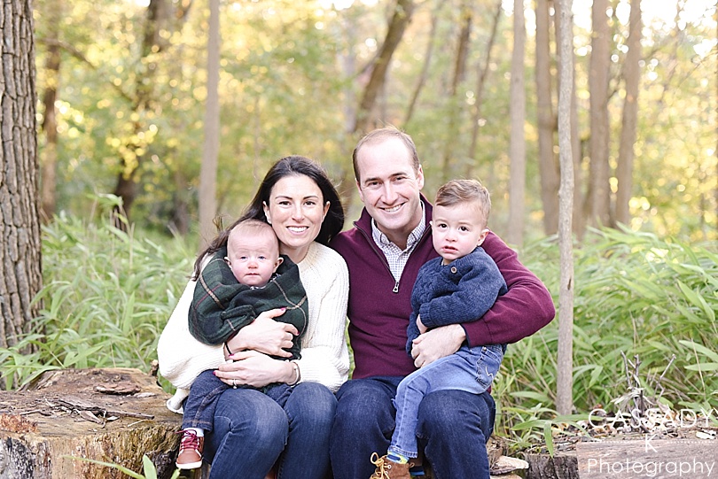 Mom and Dad with two young boys sit on logs during a Thielke Arboretum Family Session in Glen Rock, NJ