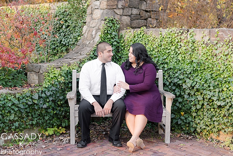 Engaged couple sits on a bench in front of ivy covered stone wall during a Cross Estate Gardens Engagement Session in Bernardsville, NJ