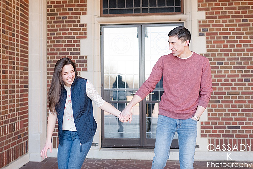 Engaged couple walking and laughing in front of red brick mansion during a Waveny House Engagement Session in New Canaan, CT