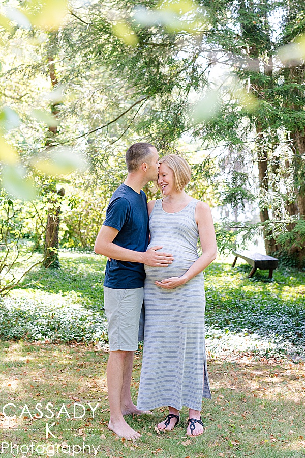 Husband kisses the forehead of his pregnant wife's head in a lush green summer outside family maternity session by Cassady K Photography