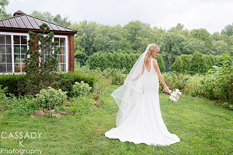 Bride shows off the back of her Sarah Seven in a garden during a Small Ninety Acres Wedding in Natirar of Peapack, NJ