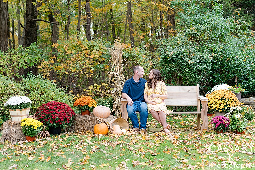 Momma-to-be and husband sit on bench, among autumn decorations at an outdoor fall maternity session in Westchester County, NY