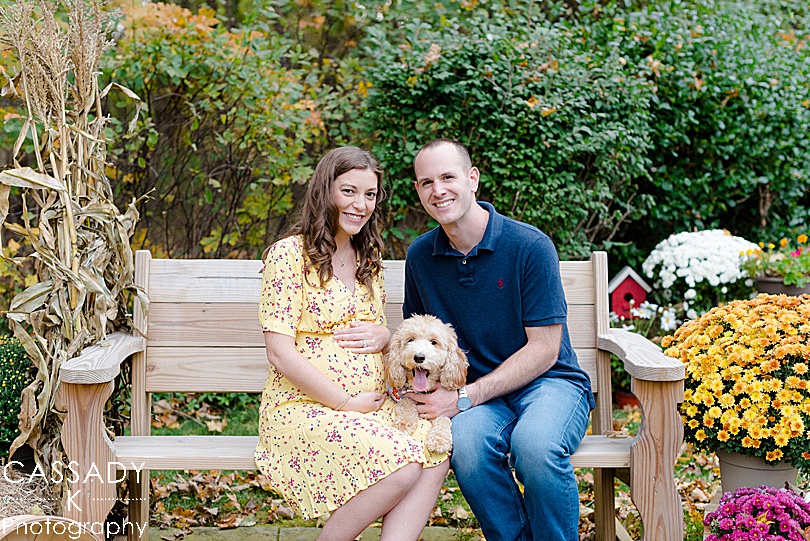 Pregnant wife and husband sit with Labradoodle outside on a bench during a fall maternity session in Westchester County, NY