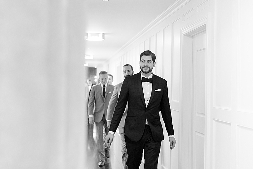Black and White of Groom walking down white paneled hall to indoor ceremony at Mansion at Natirar Wedding in Peapack, NJ