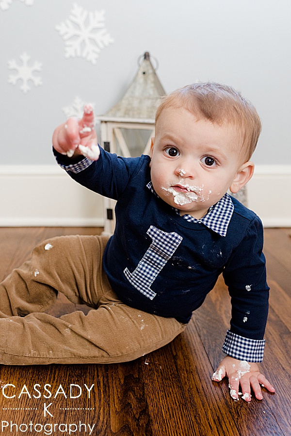 Baby boy has icing on his face from cake smash and makes a one with his finger during his at home first birthday pictures