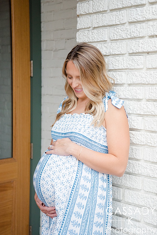 Pregnant mom looks at her bump with a baby girl in a Blue Zara dress while leaning against white brick in a Sewickley maternity session in Pittsburgh, PA