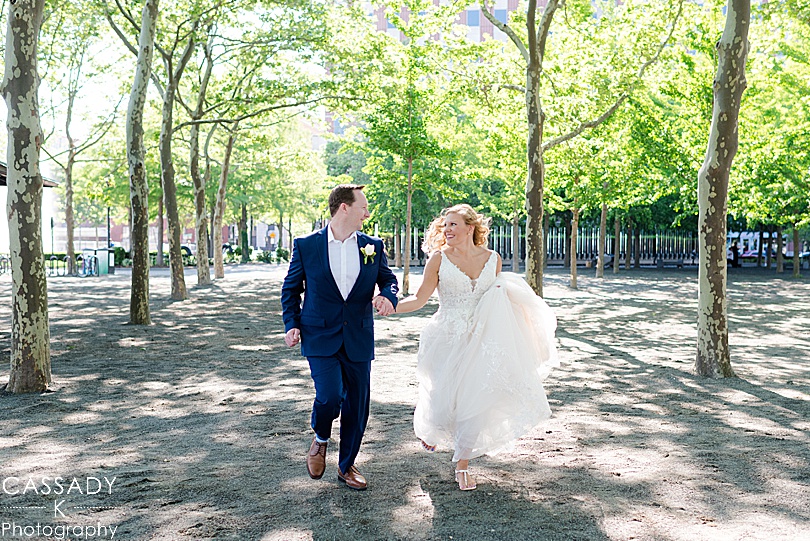 Bride and groom running under trees in Pier A in Hoboken, NJ