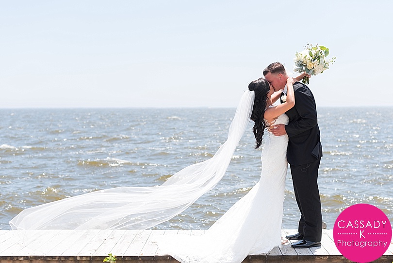 Bride and Groom kissing along the water with the cathedral length veil blowing in the water at a Bellport Country Club Wedding