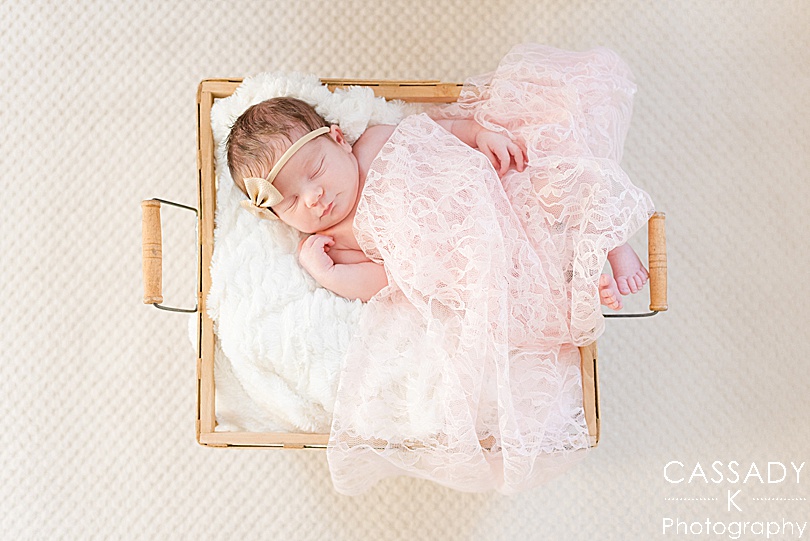 Baby girl sleeps in a tan basket with light pink lace over top of her and wearing a gold headband during a Westchester Newborn Session in Armonk, NY