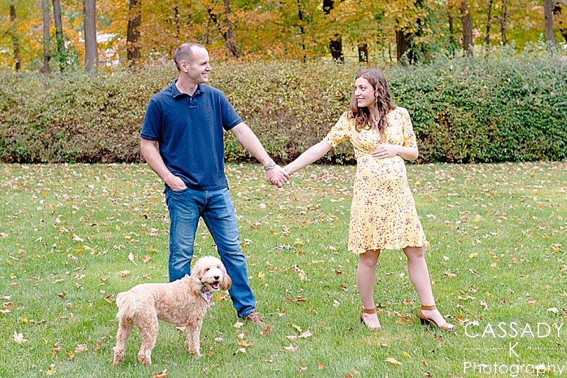 Pregnant wife and husband walk with Labradoodle outside at home during a fall maternity session in Westchester County, NY