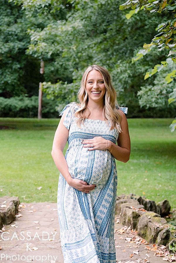 Soon to be mom walking and holding her bump in a light blue Zara sundress during an outside at home Sewickley maternity session in Pittsburgh, PA