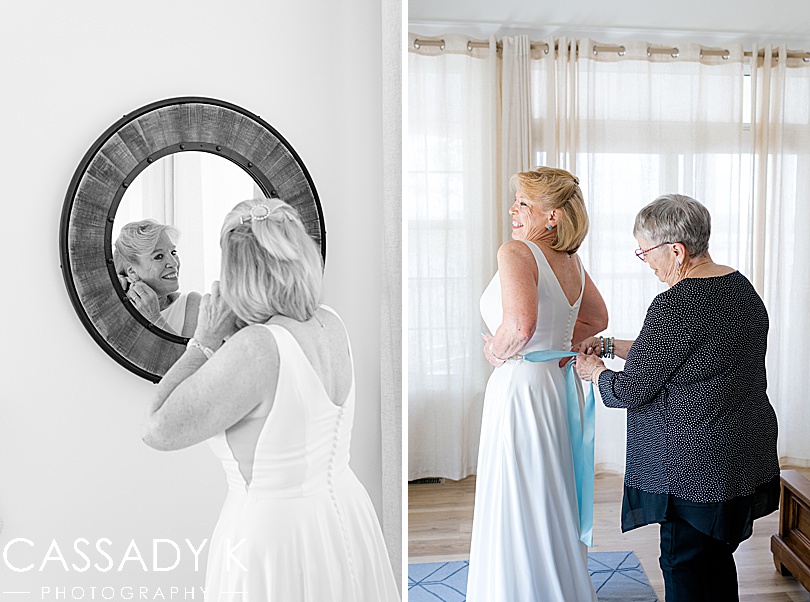 Bride putting earrings on in front of mirror and brides mother tying bow of dress