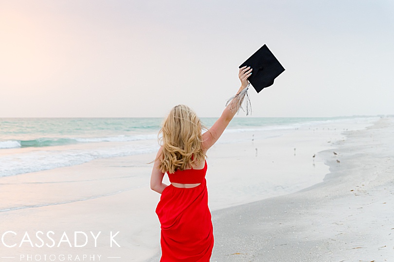 Girl in red dress holding graduation cap while on beach