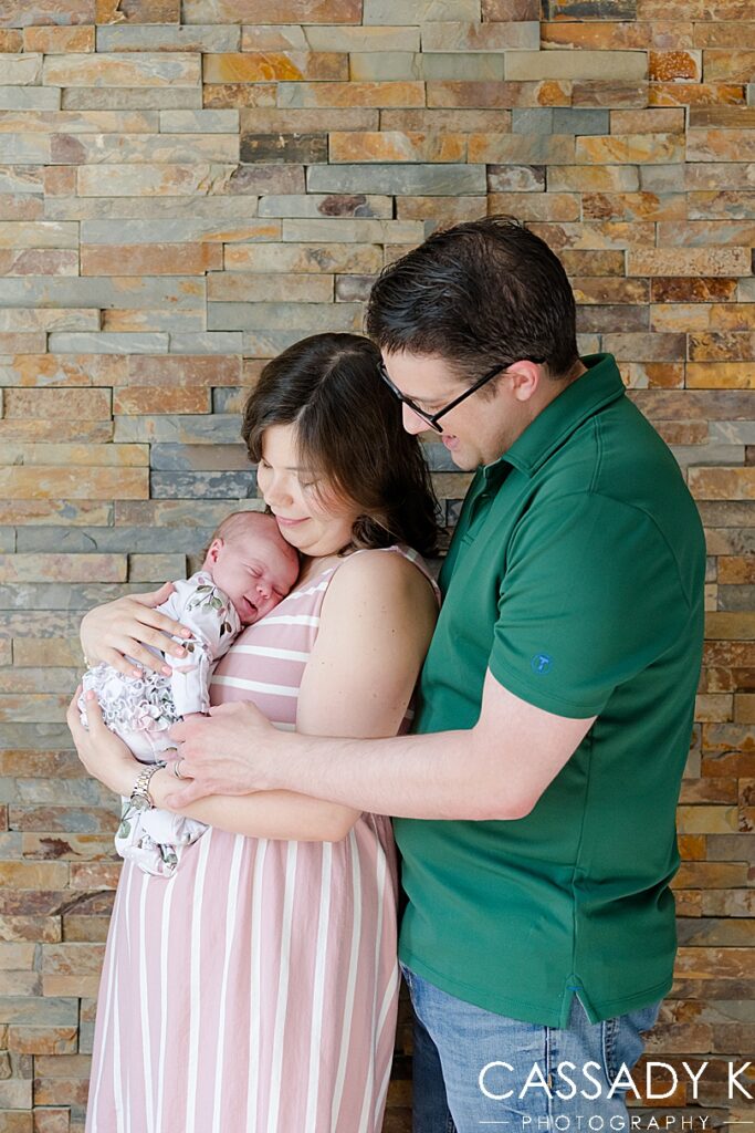 Mom and dad posing with baby girl in front of stone wall during Lansdale Lifestyle Newborn Session