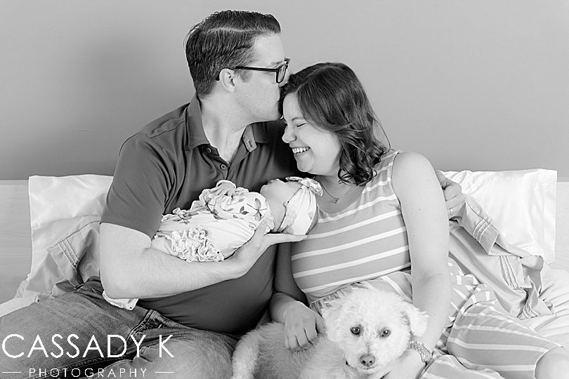 Male kissing female with baby and dog on bed during Lansdale Lifestyle Newborn Session