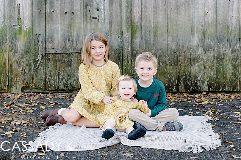 Kids sit on a blanket in front of a barn door at fall mini sesssions