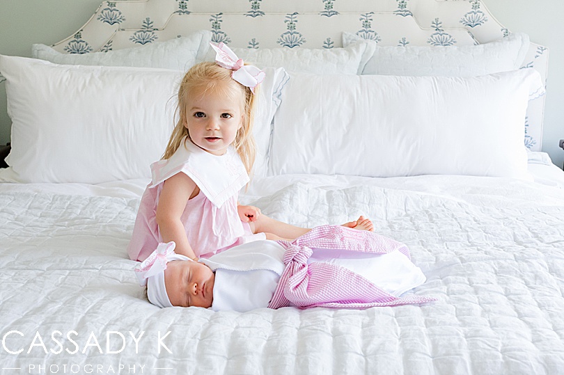 Big sister sits with her baby sister on her parents' bed during a newborn photo session.