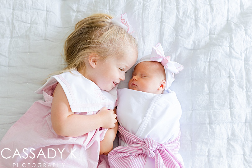 Older sister lays next to her new baby sister in their parents' bed during a newborn session outside of Philadelphia, PA